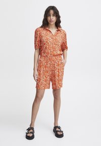 Korte mouwen oranje blouse en bijpassende shorts met een lichte bloemmotief. Voorzien van een elastische tailleband en zijzakken. Zwarte plateau sandaal.