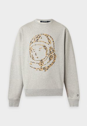 Homme en sweat-shirt gris clair avec casque d'astronaute imprimé léopard, pantalon noir à jambes larges et chaussures noires, debout sur fond blanc.