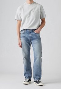 Grå bomulls T-shirt med bröstficka, ihop med ljusblå denimjeans. Bär svarta sneakers med vita sulor. Enfärgad bakgrund.