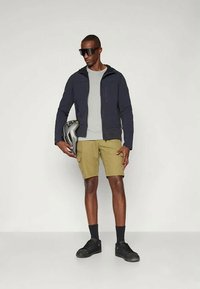 Fekete softshell dzseki, szürke póló, bézs rövidnadrág, fekete cipő, napszemüveget visel, és egy kerékpáros sisakot tart függőleges helyzetben.