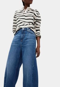 Lange blauwe denim wijde jeans gecombineerd met een crèmekleurige polo met zwarte strepen en lange mouwen. Opvallende details zijn knopen en een kraag.