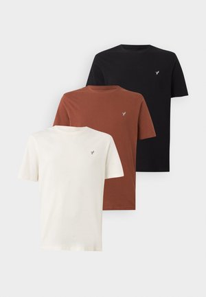 Homme debout, les mains dans les poches, portant une chemise noire à manches courtes, un pantalon gris clair et des chaussures noires sur fond blanc.