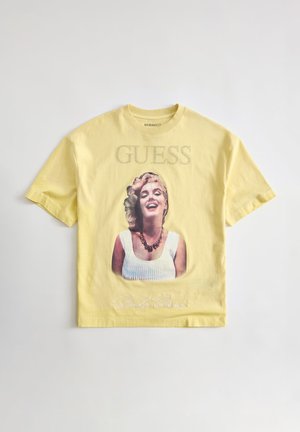T-shirt jaune à manches courtes avec une femme souriante portant un top blanc et un collier imprimés sous le texte « GUESS » sur la poitrine.