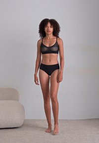 Black lace bralette and high-waisted panties, featuring a smooth texture and minimal design, model stands on a light carpeted surface.