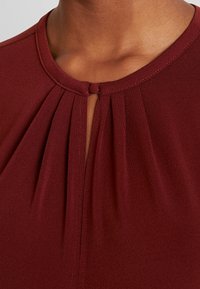 Burgundowy top z plisowanym wykończeniem, okrągłym dekoltem i detalem w kształcie łezki z przodu, wykonany z gładkiego, lekkiego materiału.