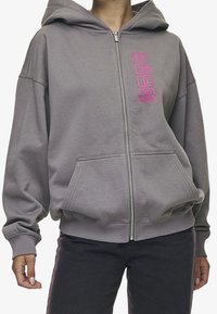 Felpa grigia con zip realizzata in morbido tessuto, dotata di cappuccio, tasche frontali e audace lettering rosa sul lato sinistro.