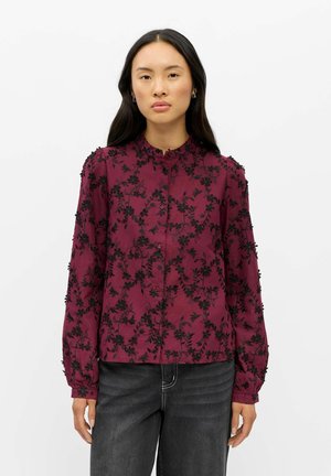 Femme aux longs cheveux foncés portant un chemisier bordeaux avec une broderie florale noire et un pantalon noir sur un fond blanc uni.