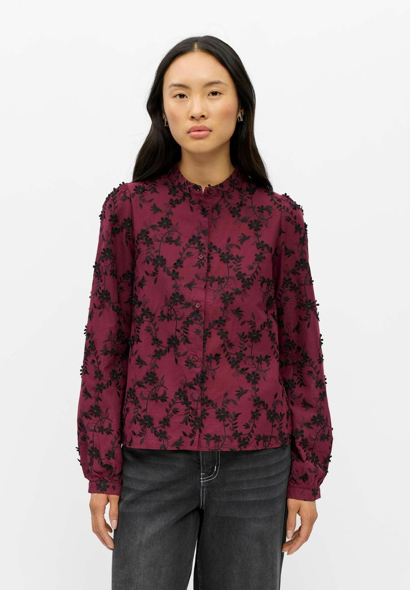 Femme aux longs cheveux foncés portant un chemisier bordeaux avec une broderie florale noire et un pantalon noir sur un fond blanc uni.