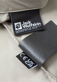 Beige stoffen label met het "Jack Wolfskin" logo, voorzien van extra labels in zwart en wit, die zorginstructies en CE-markering tonen.