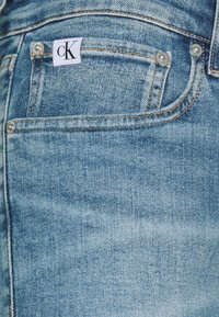 Ljust blå denimjeans med en urtvättad textur, som har en vit etikett med "cK"-logotypen och metallknappdetaljer på fickorna.