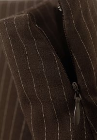 Tela marrón con finas rayas blancas, con una vista cercana de una cremallera en acabado metálico oscuro en la apertura.