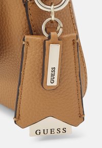 Bolso de cuero marrón claro con un acabado texturizado, que presenta un charm metálico con el logo "GUESS" y un anillo de herrajes plateados.