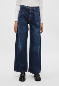 Jeans de perna larga de cintura alta em denim escuro. Apresenta pregas, dois bolsos frontais e costura em contraste. Usado com sapatos de plataforma pretos.