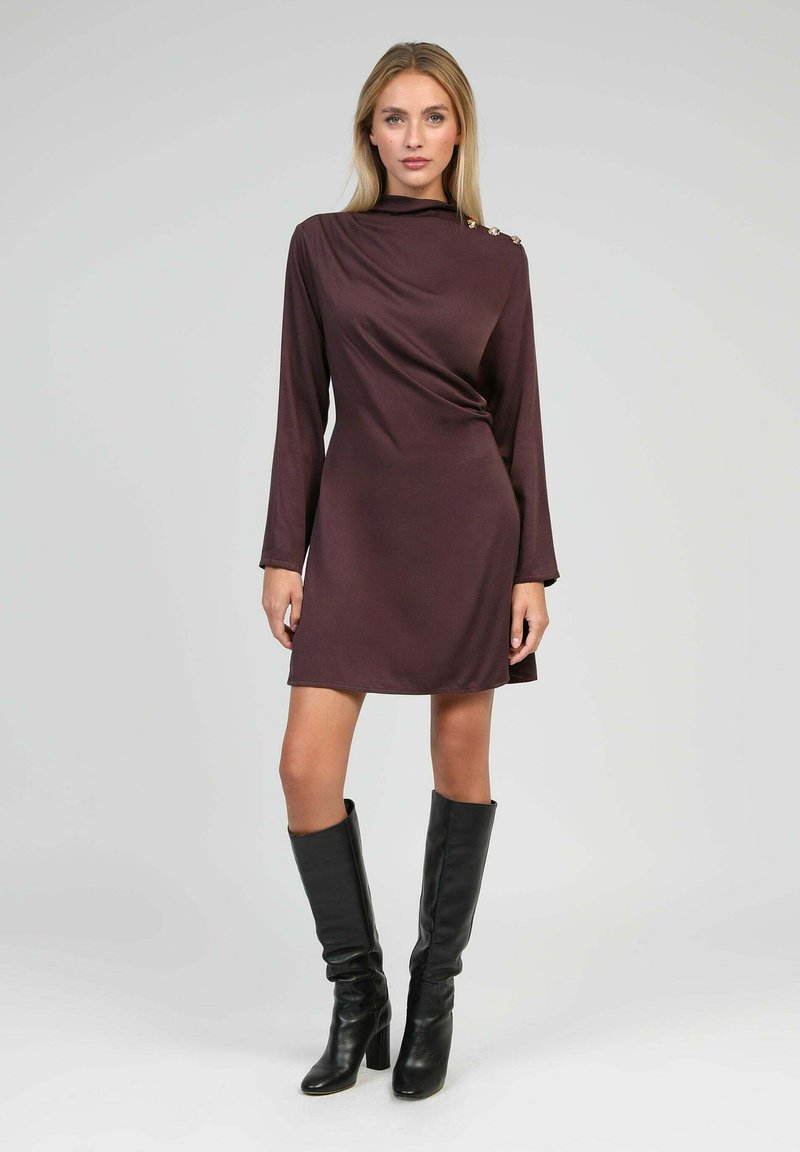 Femme portant une robe mini à manches longues marron foncé avec des boutons dorés sur les épaules et des bottes noires à talons hauts jusqu'aux genoux, se tenant devant un fond uni.