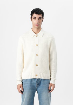 Cremefarbener Strickcardigan mit kragenförmigem Ausschnitt, durchgehender Knopfleiste, gerippten Bündchen und einem strukturierten Muster. Getragen über blauen Jeans.