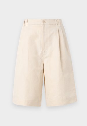 Nainen pukeutunut beigeen ribbineuleeseen, off-white polvipitkiin shortseihin, joissa musta vyö, pitää taupen väristä käsilaukkua ja mustia tasapohjaisia kenkiä, joissa on remmit.