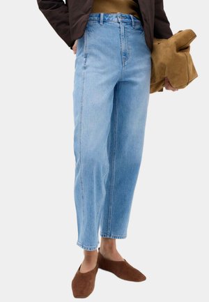 Frau trägt hellblaue High-Waist-Jeans, braune Slip-On-Schuhe, hält eine beige Wildledertasche und trägt eine dunkle Jacke und ein grünes Oberteil.