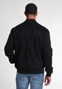 Jacket bomber din suede negru, cu un tiv și mansete ribbed, având o textură netedă și un design simplu pe spate. Purtați cu blugi albastri deschis.