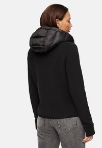 Zwarte hoodie met een gevoerde zwarte capuchon, zachte textuur en aansluitend ontwerp. Heeft naadloze stiksels en geribbeld elastiek aan de mouwen, gedragen met grijze jeans.