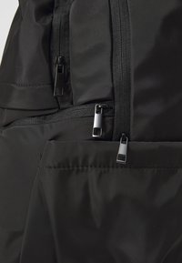 Zaino in nylon nero con texture liscia, dotato di molteplici tasche con zip, cerniere metalliche e dettagli cuciti.