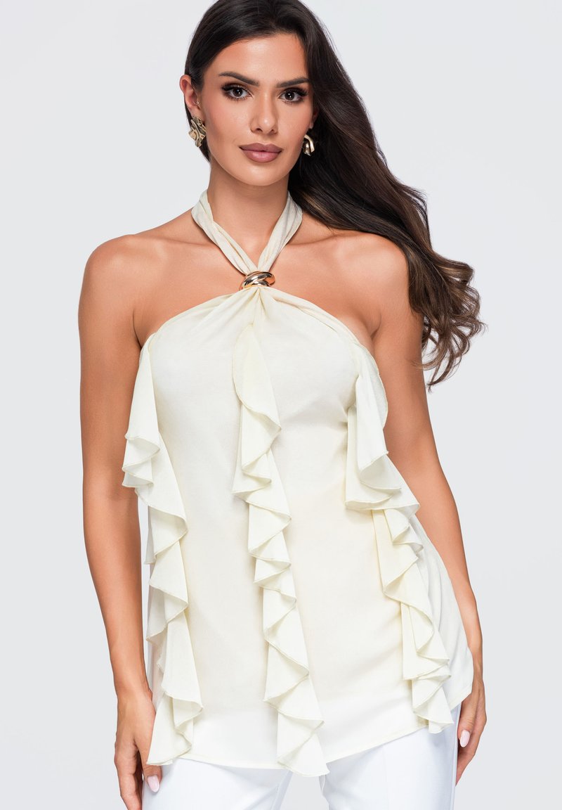 Femme aux longs cheveux foncés portant un top blanc sans manches à col licou avec des détails à volants et un anneau métallique au niveau du décolleté, sur un fond uni.