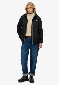 Sort vandtæt jakke med hætte, kontrastlynlås og logo; båret over en let striktrøje og blå denimjeans, parret med sorte støvler.