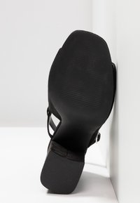 Chaussure à talon noire avec bout carré et bride à la cheville, semelle et talon texturés pour l'adhérence, placée devant un mur et un sol blancs.