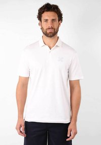 Chemise polo blanche avec un col classique et des manches courtes. Présente un logo floral bleu subtil sur la poitrine gauche. Texture lisse.