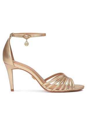 Goldene High-Heel-Sandale mit offenen Zehen, mehreren vorderen Riemen und einem dünnen Knöchelriemen mit einem kleinen runden Anhänger.