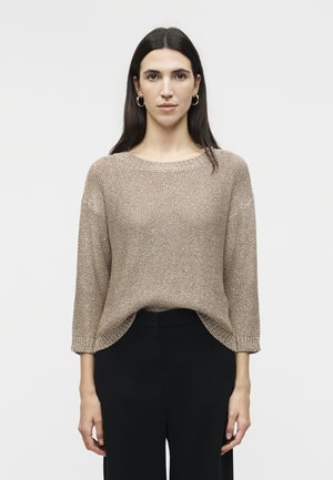 Hosszú sötét hajú nő, arany karika fülbevalóval, laza bézs kötött pulóverrel és fekete nadrággal, fehér háttér előtt áll.