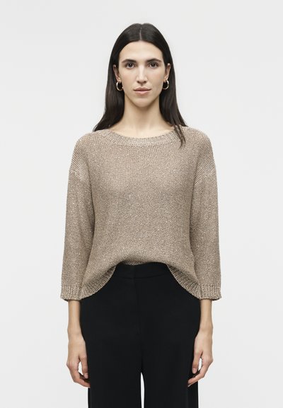 Femme aux longs cheveux noirs portant des boucles d'oreilles créoles en or, un pull beige en maille ample et un pantalon noir, debout sur un fond blanc.