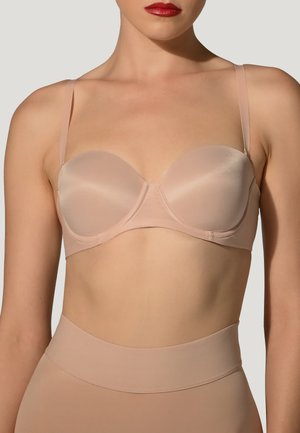 Kvinde iført en nude strapless bh med tynde aftagelige stropper og matchende højtaljet sømløst shapewear mod en ensfarvet baggrund.