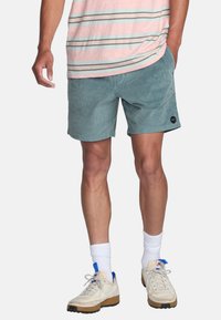 Shorts bleu-vert clair avec une texture douce, dotés de poches latérales et d'une simple étiquette noire. Associés à une chemise rayée rose et des baskets.