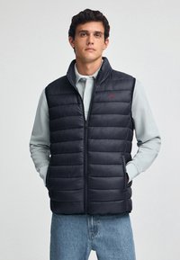 Granatowa kamizelka puchowa z pikowanym wzorem, przednim zamkiem błyskawicznym, wysokim kołnierzem i czerwonym akcentem w postaci logo, noszona na jasnoszarym długim rękawie i dżinsach.