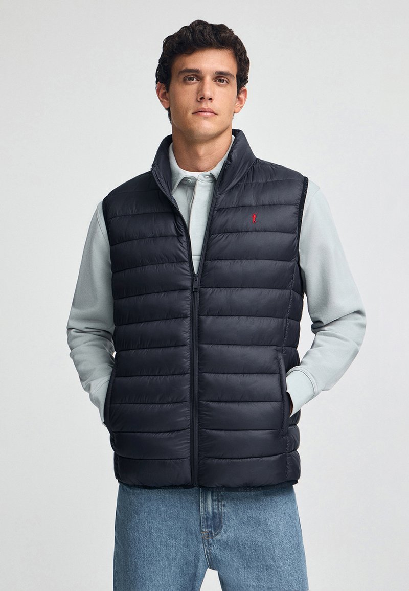 Granatowa kamizelka puchowa z pikowanym wzorem, przednim zamkiem błyskawicznym, wysokim kołnierzem i czerwonym akcentem w postaci logo, noszona na jasnoszarym długim rękawie i dżinsach.