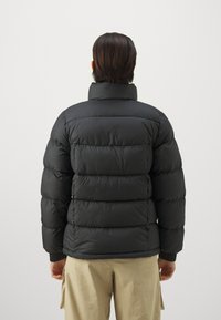 Czarna kurtka puchowa z pionowymi przeszyciami i wysokim kołnierzem. Ma prążkowane mankiety. Nosi ją osoba, prezentując widok z tyłu na neutralnym tle.