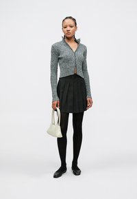 Sivi kratki pleteni kardigan s ovratnikom, uparen s crnom plisiranom mini suknjom i jednobojnim crnim čarapama, držeći malu bijelu torbicu.