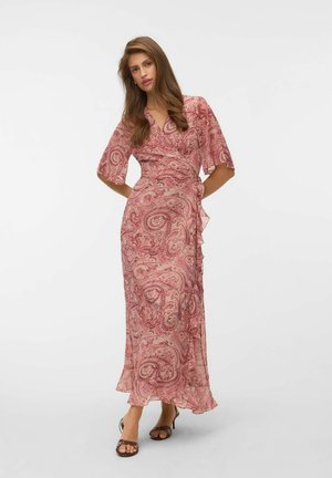 Femme debout dans une robe portefeuille rose à motif cachemire avec manches jusqu'au coude et sandales à talons noires à brides sur fond blanc.