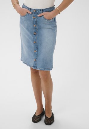 Naine, kes kannab sinist nööpidega teksaseelik, käed taskutes, kuldsete käevõrude ja mustade punutud tasaste kingadega, seisab valgel taustal.