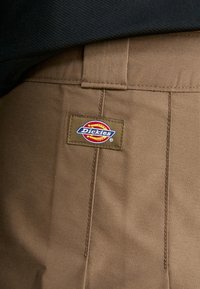Pantaloni marroni Dickies con una toppa rettangolare con testo multicolore e un design circolare. Il materiale sembra durevole con tessuto testurizzato.