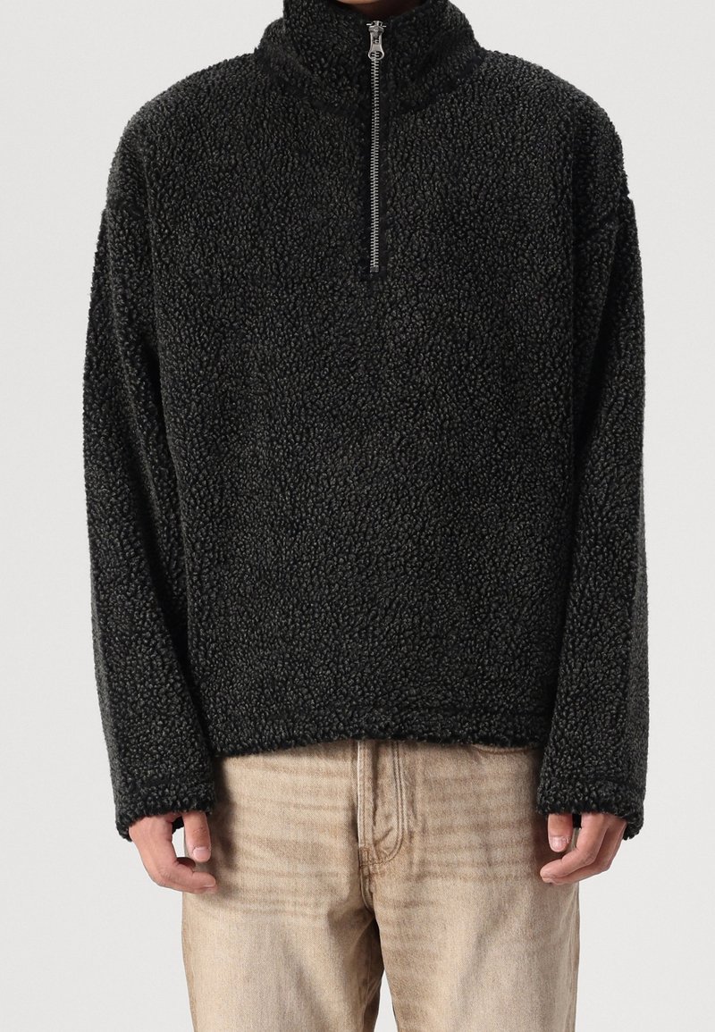 Suéter de forro polar negro con cuello alto, cierre de cremallera en la parte delantera y mangas largas. Superficie texturizada con una apariencia suave y esponjosa. Corte casual.