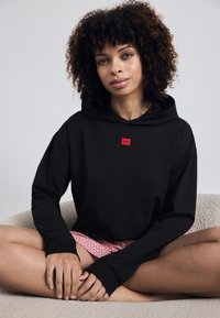 Fekete kapucnis pulóver piros logópatch-el, laza szabással, hosszú ujjakkal és elülső kenguru zsebével. Texturált anyag, egyszerű dizájn.