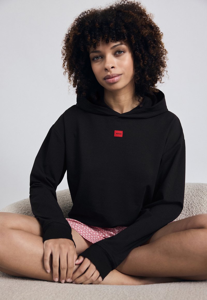 Fekete kapucnis pulóver piros logópatch-el, laza szabással, hosszú ujjakkal és elülső kenguru zsebével. Texturált anyag, egyszerű dizájn.