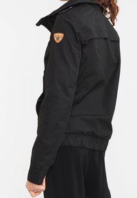 Svart, åtsittande jacka gjord av slitstarkt tyg, med dragkedja fram, elastiska ärmslut och en logotypapplikation på axeln.