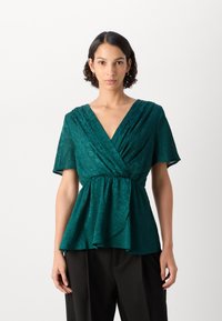 Blusa verde acqua con scollo a V e maniche corte. Il tessuto presenta un delicato motivo a trama, cinte in vita, che si apre in un design a peplum.