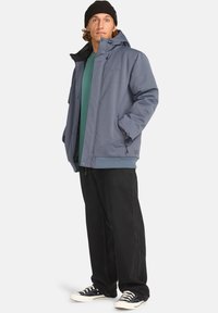 Grå isolerad jacka med huva, blå ribbade muddar och sidfickor; bärs över en grön skjorta med svarta löst sittande byxor och sneakers.