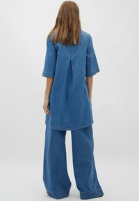 Denim tuniek met een rugplooitje, korte mouwen en een losse pasvorm, gecombineerd met wijde denim broekspijpen. Beide in een middenblauwe was.