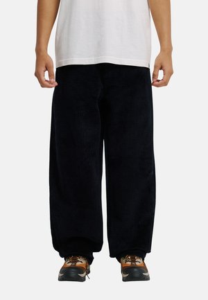 Persoon draagt zwarte, wijde broek, bruin-oranje sneakers en een wit T-shirt, staat met ontspannen handen langs het lichaam.
