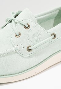 Chaussure bateau en daim vert clair avec œillets métalliques ronds, lacets assortis, coutures moc toe, et semelle en caoutchouc blanche.