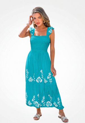 Mujer con vestido sin mangas color turquesa con estampados florales blancos, con pendientes de perlas y sandalias, de pie contra un fondo claro.